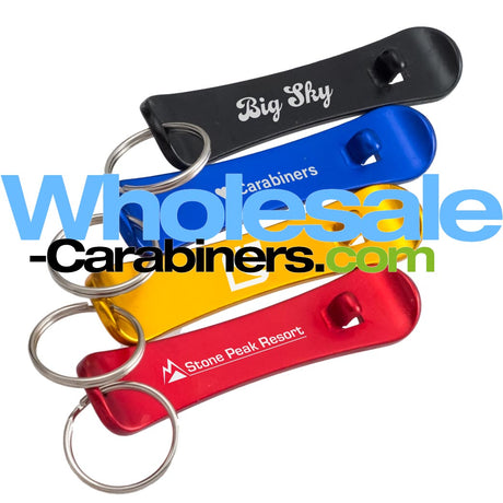 Alternate view of four aluminum bottle opener keychains shaped like snowboards. The snowboards are black, blue, gold, and red and each shows an example engraved logo. Overlayed with a wholesale-carabiners.com watermark.